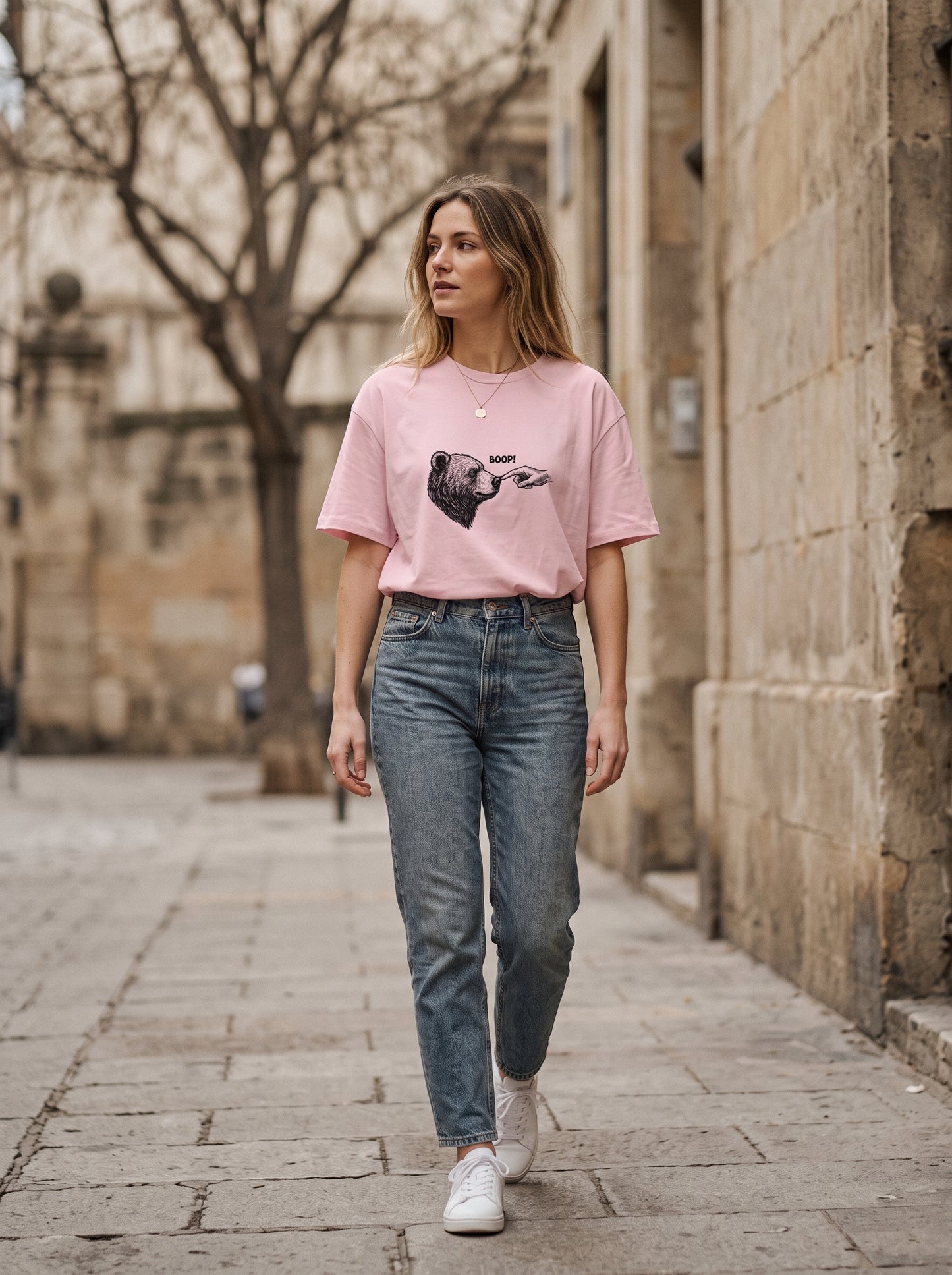 A woman walking down a stone street wearing a light pink graphic t-shirt, blue jeans, and white sneakers.